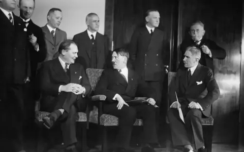 Am 30. Januar 1933: Hitler und seine Gefolgschaft, eine Gruppe von Männern in Anzügen, sitzen und stehen in einem Raum.