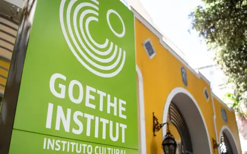 Grünes Schild mit weißem Logo und Schriftzug "GOETHE INSTITUT" vor einem gelben Gebäude.