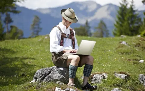 Mann in Lederhose und Hut arbeitet auf einem Laptop in den Bergen.