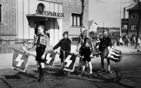 Jungen in Uniformen tragen Fahnen mit Hakenkreuzen und Trommeln. Ein junger Mann, der an Hitler erinnert, marschiert mit ihnen.