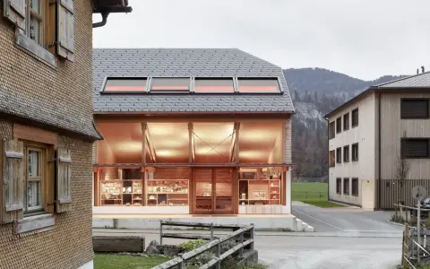 Neues Bauen: Moderne Architektur trifft auf traditionelle Schindelfassade. Ein hell erleuchtetes Glasgebäude mit Ausstellungsstücken steht neben einem Holzhaus.