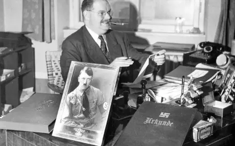 Philipp Auerbach sitzt an seinem Schreibtisch, eine Pfeife im Mund, und betrachtet ein Bild von Adolf Hitler. Daneben liegt eine Urkunde.