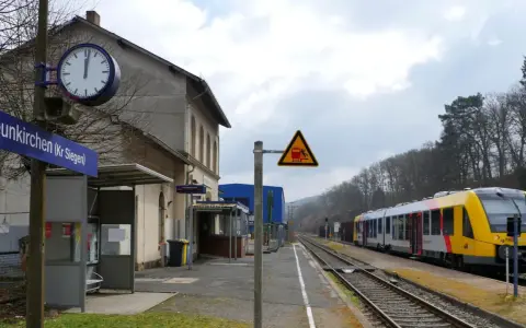 Bahnhof Neunkirchen (Kr Siegen) mit gelb-silbernem Zug auf den Gleisen. Ein Warnschild zeigt einen Zug und eine stürzende Person.