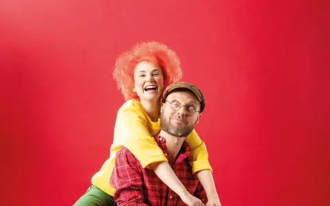 Ein Mann mit Brille und Kappe, der wie Meister Eder aussieht, trägt eine Frau mit feuerroter Perücke auf dem Rücken. Sie lachen ausgelassen.