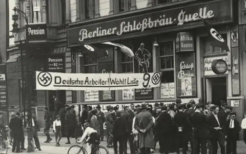 Menschenmenge vor einem Gebäude mit der Aufschrift "Berliner Schloßbräu-Quelle". Ein Banner verkündet "Deutsche Arbeiter! Wählt Liste 9". Hakenkreuzfahnen sind sichtbar, was auf die Zeit vor dem NSDAP Verbot hinweist.