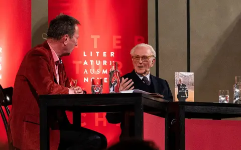 Zwei Männer im Gespräch, einer mit Buch "Franz von Bayern". Einblicke in die bayerische Geschichte, vielleicht aus dem Jahr 794.