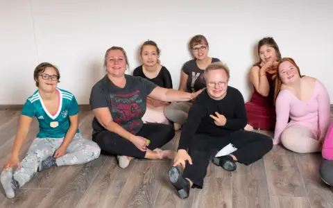 Gruppe von Menschen mit Down-Syndrom und einer Betreuerin sitzen auf dem Boden.
