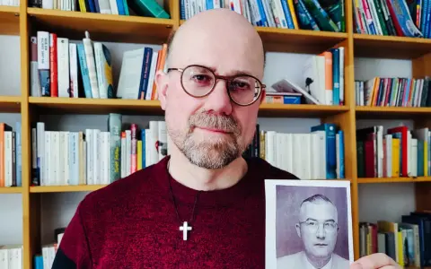Mann mit Brille hält Foto eines Mannes in Uniform. Im Hintergrund Bücherregale. Das Foto zeigt einen Mann, der der SS Schutzstaffel angehörte.