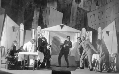 Szene einer Theateraufführung mit expressionistischem Bühnenbild. Menschen agieren auf der Bühne, als würden sie **trommeln in der Nacht**.