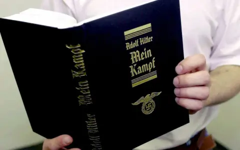 Eine Person hält ein Buch mit dem Titel "Mein Kampf" von Adolf Hitler. Das Buch hat einen schwarzen Einband mit goldener Schrift und einem Hakenkreuz-Adler-Symbol.