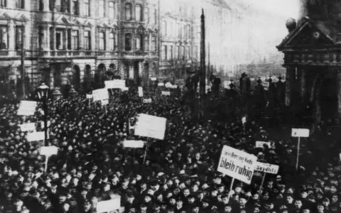 Massen demonstrieren in der Revolution 1918. Schilder mit Aufschriften wie "Seydlitz" und "Bleib ruhig" werden hochgehalten.