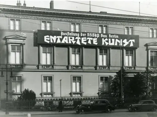 Große Banner verkünden die "Ausstellung der NSDAP Gau Berlin ENTTARTETE KUNST" an einem Gebäude. Autos und Fahrräder stehen davor.