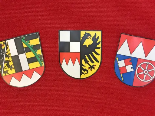 Drei Wappen auf rotem Grund: links ein Löwe, Mitte ein Adler, rechts ein Fähnlein und ein Rad. Sie repräsentieren bayerische Regierungsbezirke.