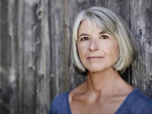 Vroni Well, eine Frau mit grauem Haar und einem warmen Blick, lehnt an einer rustikalen Holzwand.