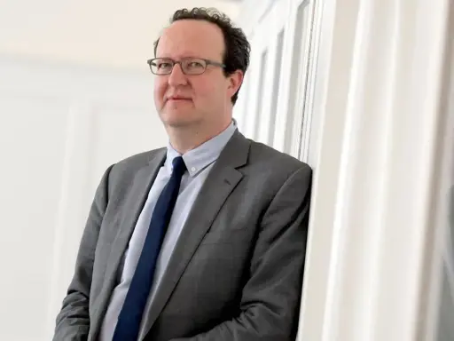 Hans Ottomeyer, ein Mann mit Brille im grauen Anzug, lehnt an einer weißen Wand.