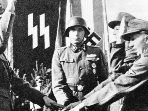 Ein SS-Offizier im Helm steht vor einer Fahne mit SS-Runen. Mehrere junge Männer in Uniformen sind zu sehen.