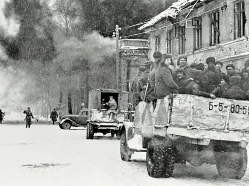 Soldaten der Roten Armee 1945 fahren in einem LKW durch eine verschneite Stadt. Rauch steigt aus einem zerstörten Gebäude auf.