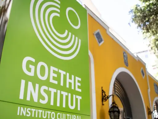 Grünes Schild mit weißem Logo und Schriftzug "GOETHE INSTITUT" vor einem gelben Gebäude.