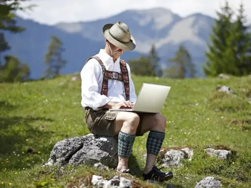 Mann in Lederhose und Hut arbeitet auf einem Laptop in den Bergen.