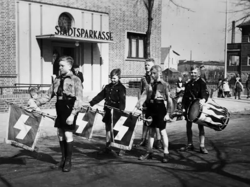 Jungen in Uniformen tragen Fahnen mit Hakenkreuzen und Trommeln. Ein junger Mann, der an Hitler erinnert, marschiert mit ihnen.