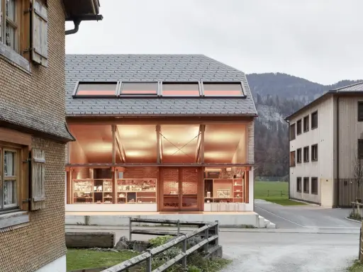 Neues Bauen: Moderne Architektur trifft auf traditionelle Schindelfassade. Ein hell erleuchtetes Glasgebäude mit Ausstellungsstücken steht neben einem Holzhaus.