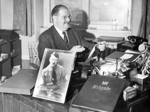 Philipp Auerbach sitzt an seinem Schreibtisch, eine Pfeife im Mund, und betrachtet ein Bild von Adolf Hitler. Daneben liegt eine Urkunde.