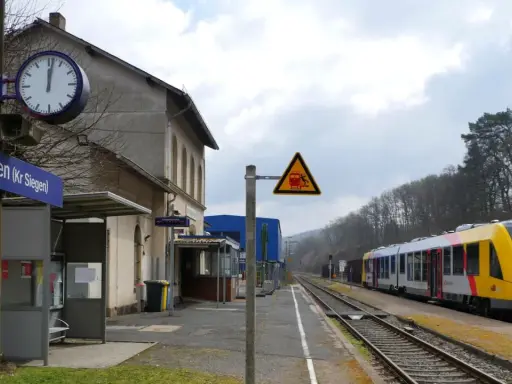 Bahnhof Neunkirchen (Kr Siegen) mit gelb-silbernem Zug auf den Gleisen. Ein Warnschild zeigt einen Zug und eine stürzende Person.