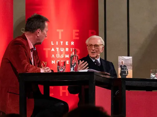Zwei Männer im Gespräch, einer mit Buch "Franz von Bayern". Einblicke in die bayerische Geschichte, vielleicht aus dem Jahr 794.