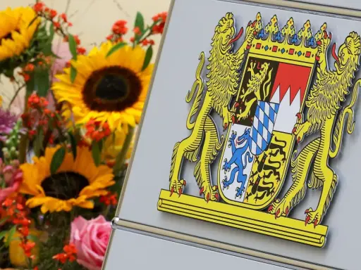 Das Staatswappen Bayerns, flankiert von Löwen, steht stolz neben einem bunten Blumenstrauß mit Sonnenblumen und Rosen.
