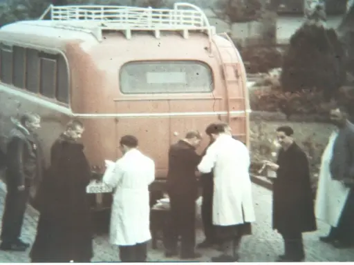 Männer in weißen Kitteln und dunklen Mänteln stehen vor einem roten Bus. Ein Kennzeichen ist sichtbar. Die Szene könnte mit der NS-Euthanasie in Verbindung stehen.