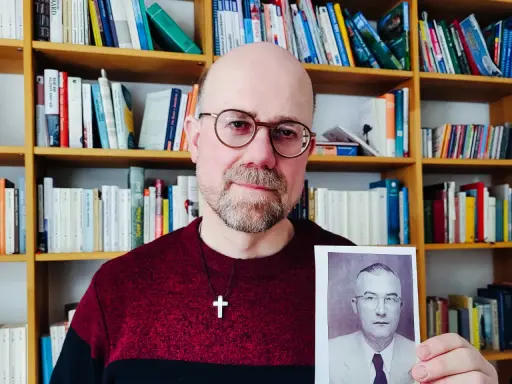 Mann mit Brille hält Foto eines Mannes in Uniform. Im Hintergrund Bücherregale. Das Foto zeigt einen Mann, der der SS Schutzstaffel angehörte.