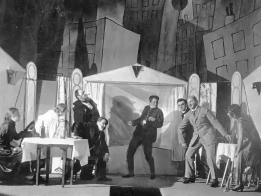 Szene einer Theateraufführung mit expressionistischem Bühnenbild. Menschen agieren auf der Bühne, als würden sie **trommeln in der Nacht**.