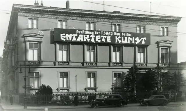 Große Banner verkünden die "Ausstellung der NSDAP Gau Berlin ENTTARTETE KUNST" an einem Gebäude. Autos und Fahrräder stehen davor.