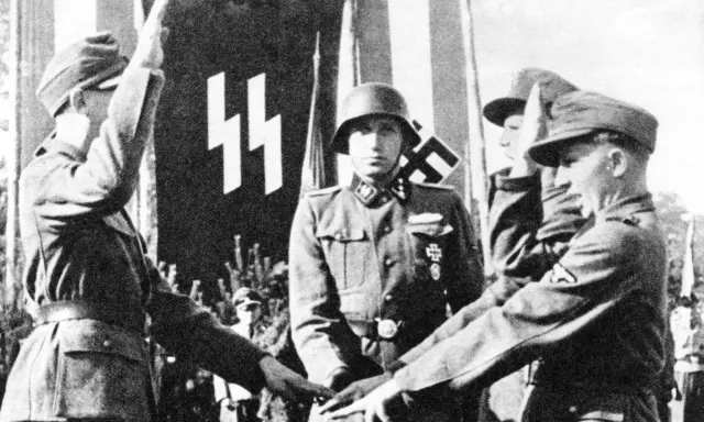 Mehrere SS-Soldaten stehen vor einer Fahne mit SS-Runen. Einer hebt den Arm zum Gruß.