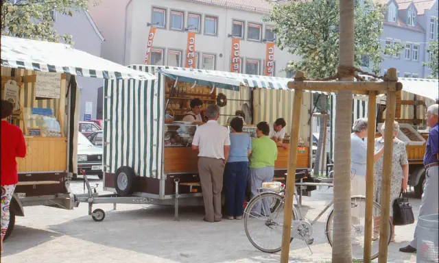 Marktstände in Neukirchen Balbini mit Brot und Butter. Menschen kaufen ein, ein Fahrrad steht bereit.