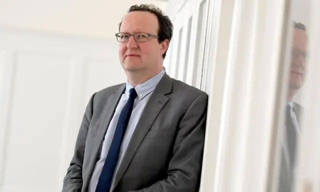 Hans Ottomeyer, ein Mann mit Brille im grauen Anzug, lehnt an einer weißen Wand.