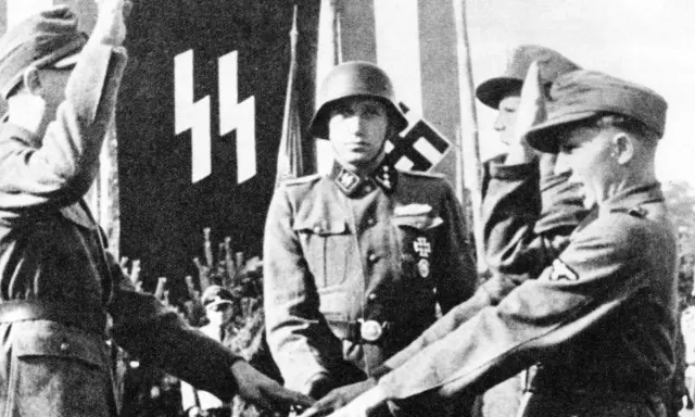 Ein SS-Offizier im Helm steht vor einer Fahne mit SS-Runen. Mehrere junge Männer in Uniformen sind zu sehen.