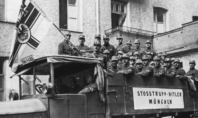 Männer in Uniformen, die an den Hitler-Putsch erinnern, stehen auf einem Lastwagen mit einer Fahne.