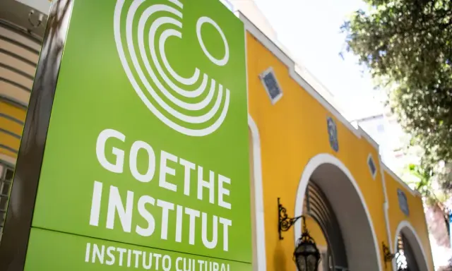 Grünes Schild mit weißem Logo und Schriftzug "GOETHE INSTITUT" vor einem gelben Gebäude.
