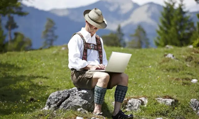 Mann in Lederhose und Hut arbeitet auf einem Laptop in den Bergen.
