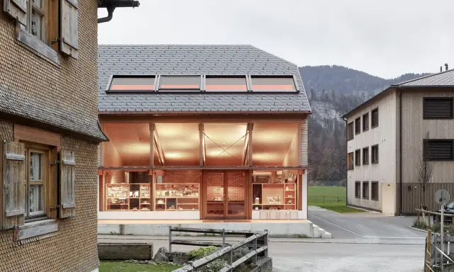 Neues Bauen: Moderne Architektur trifft auf traditionelle Schindelfassade. Ein hell erleuchtetes Glasgebäude mit Ausstellungsstücken steht neben einem Holzhaus.