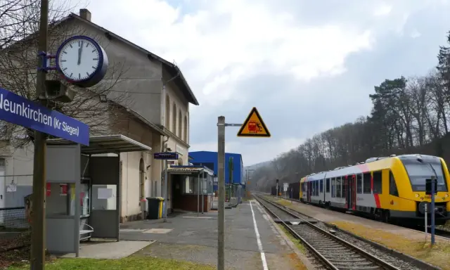 Bahnhof Neunkirchen (Kr Siegen) mit gelb-silbernem Zug auf den Gleisen. Ein Warnschild zeigt einen Zug und eine stürzende Person.