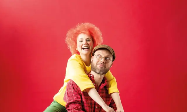 Ein Mann mit Brille und Kappe, der wie Meister Eder aussieht, trägt eine Frau mit feuerroter Perücke auf dem Rücken. Sie lachen ausgelassen.