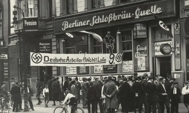 Menschenmenge vor einem Gebäude mit der Aufschrift "Berliner Schloßbräu-Quelle". Ein Banner verkündet "Deutsche Arbeiter! Wählt Liste 9". Hakenkreuzfahnen sind sichtbar, was auf die Zeit vor dem NSDAP Verbot hinweist.