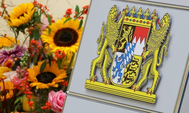 Das Staatswappen Bayerns, flankiert von Löwen, steht stolz neben einem bunten Blumenstrauß mit Sonnenblumen und Rosen.