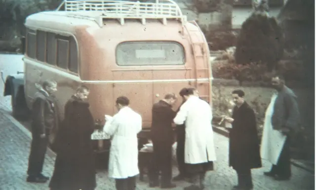 Männer in weißen Kitteln und dunklen Mänteln stehen vor einem roten Bus. Ein Kennzeichen ist sichtbar. Die Szene könnte mit der NS-Euthanasie in Verbindung stehen.
