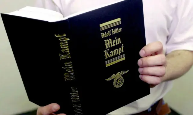 Eine Person hält ein Buch mit dem Titel "Mein Kampf" von Adolf Hitler. Das Buch hat einen schwarzen Einband mit goldener Schrift und einem Hakenkreuz-Adler-Symbol.