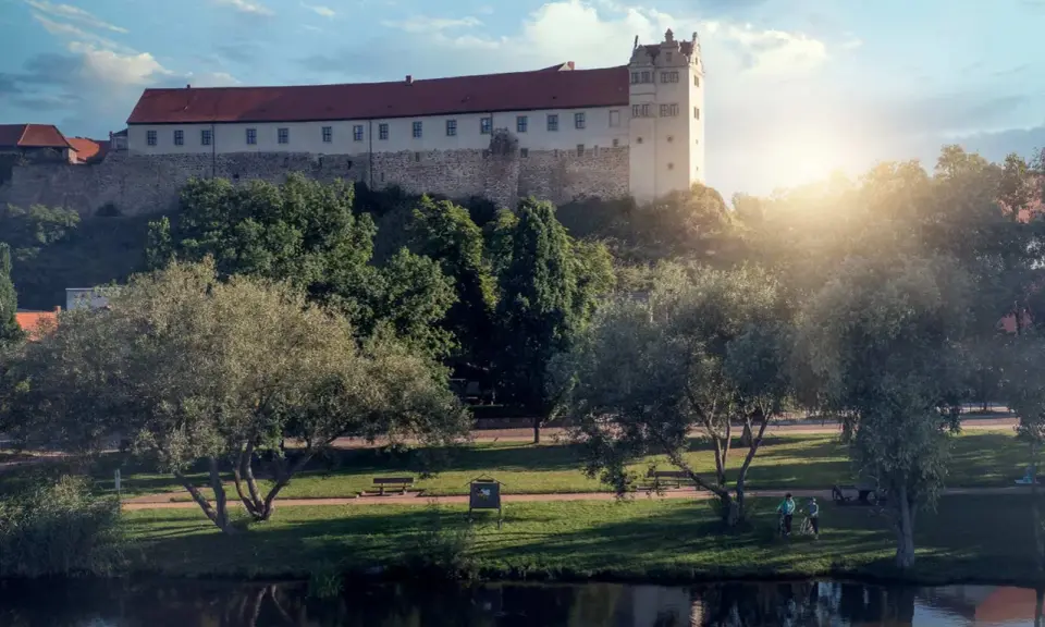 Schloss hinter Bäumen, ein Fluss im Vordergrund. Ein friedlicher Tag, wie ihn die Wettiner heute genießen könnten.