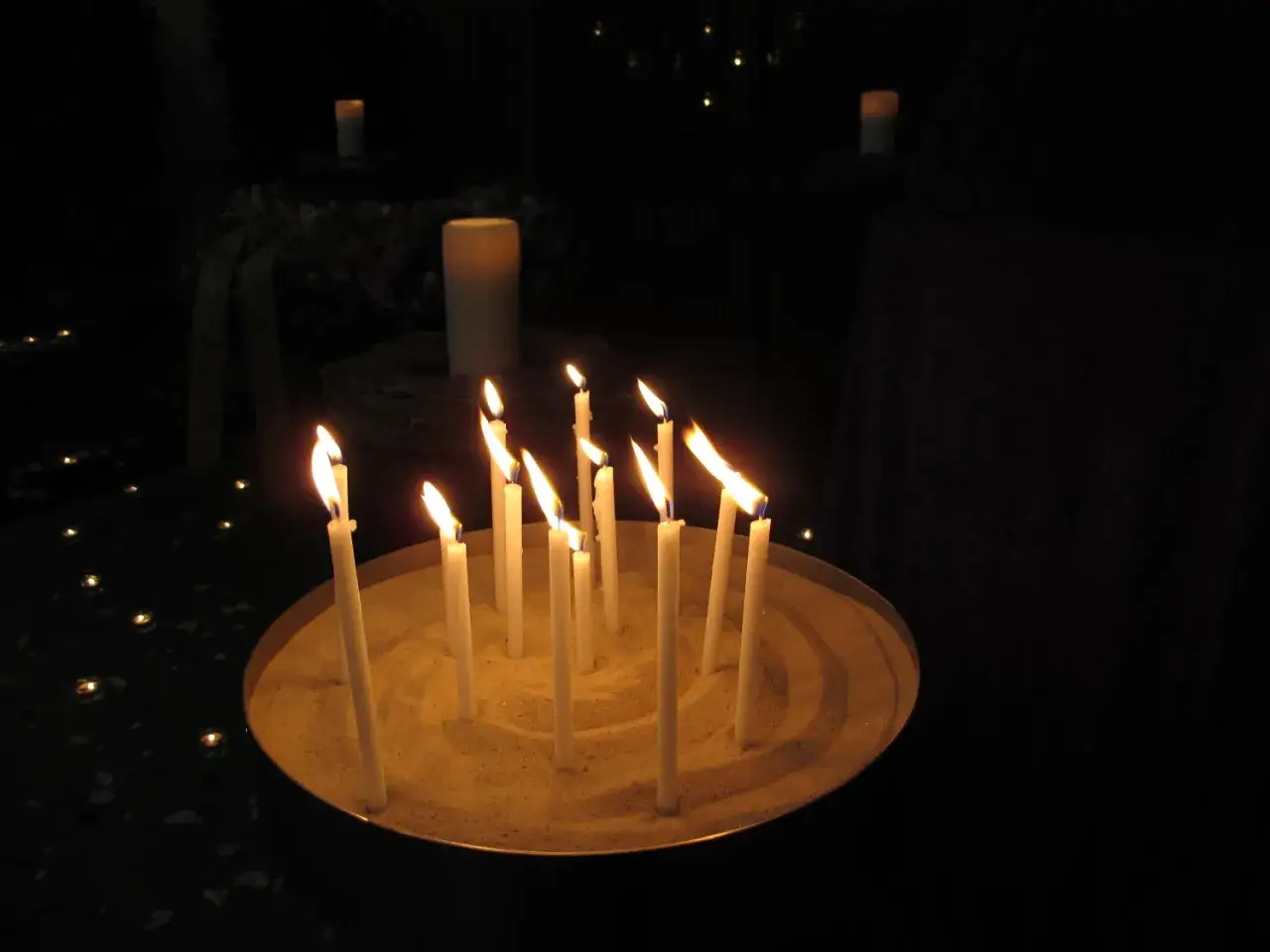 Brennende Kerzen in einer Schale mit Sand, ein Symbol f&uuml;r Bestattungsrituale im Christentum. Ein Kranz und eine S&auml;ule sind im Hintergrund zu sehen.