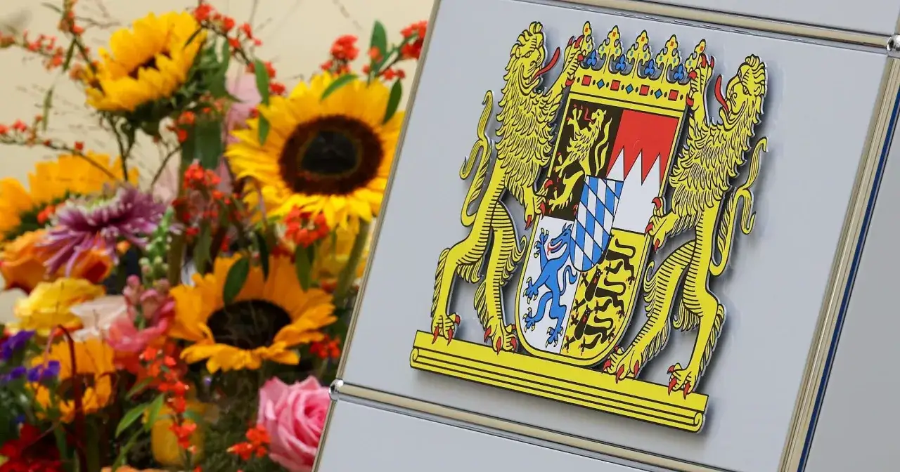 Das Staatswappen Bayerns, flankiert von Löwen, steht stolz neben einem bunten Blumenstrauß mit Sonnenblumen und Rosen.